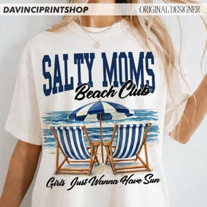 Op de afbeelding: Cr&egrave;mekleurig T-shirt met de tekst "SALTY MOMS Beach Club" in marineblauw en zwart. De afbeelding toont een strandsc&egrave;ne met blauw-wit gestreepte stoelen, een parasol en de woorden "Girls Just Wanna Have Sun".