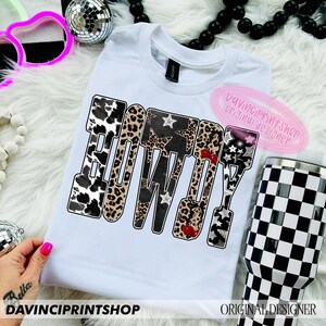 May include: White t-shirt with the word "BOWDY" in a bold, patterned design. The letters feature cow print, leopard print, stars, and cherries. A black and white checkered tumbler is visible.