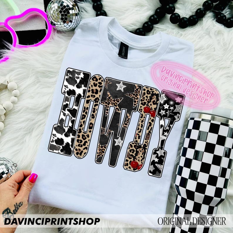 May include: White t-shirt with the word "BOWDY" in a bold, patterned design. The letters feature cow print, leopard print, stars, and cherries. A black and white checkered tumbler is visible.