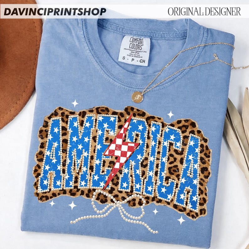 May include: A light blue t-shirt with the word "AMERICA" in blue stars, outlined with white gems, and a red and white checkered lightning bolt. The word is set against a leopard print background. A gold necklace and a pearl bow are also visible.