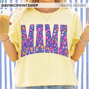 May include: A light yellow t-shirt with the word "MAMA" in large, floral-filled letters. The flowers are in shades of purple, pink, orange, and blue. The shirt has short sleeves and a crew neck. The text "DAVINCIPRINTSHOP" and "ORIGINAL DESIGNER" are at the top.