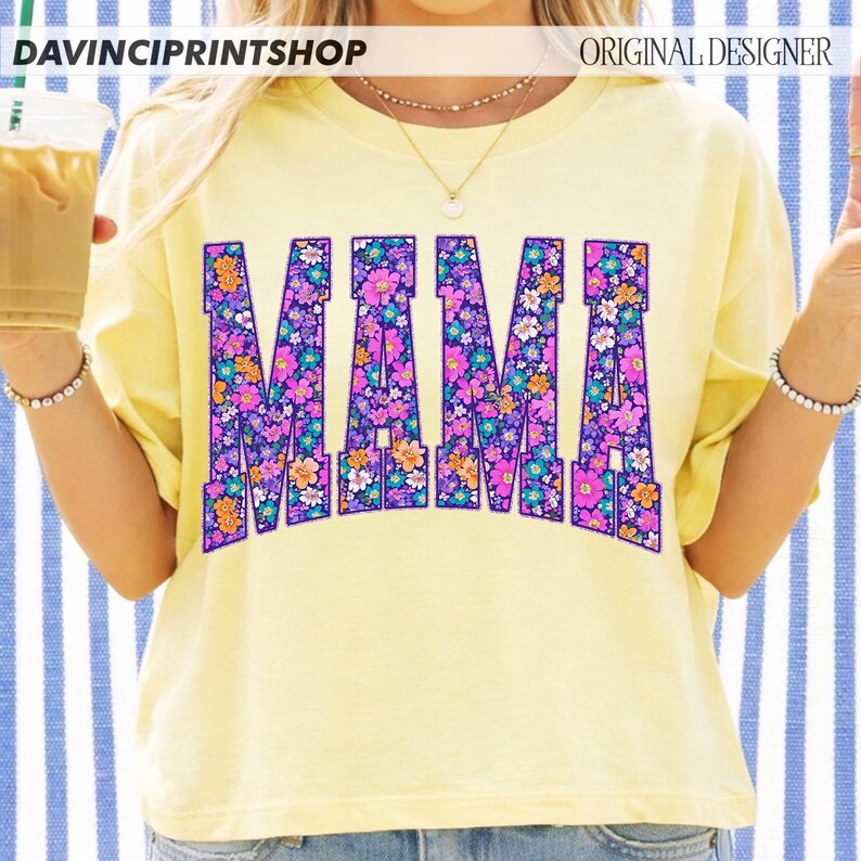 May include: A light yellow t-shirt with the word "MAMA" in large, floral-filled letters. The flowers are in shades of purple, pink, orange, and blue. The shirt has short sleeves and a crew neck. The text "DAVINCIPRINTSHOP" and "ORIGINAL DESIGNER" are at the top.