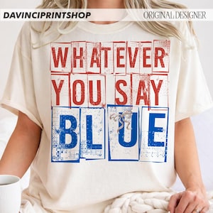 以下が含まれることがあります： クリーム色のTシャツ。赤と青のブロック文字で「WHATEVER YOU SAY BLUE」と書かれています。デザインには小さなリボンが付いています。カジュアルで快適なスタイルです。