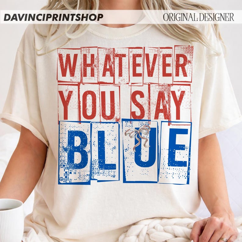 以下が含まれることがあります： クリーム色のTシャツ。赤と青のブロック文字で「WHATEVER YOU SAY BLUE」と書かれています。デザインには小さなリボンが付いています。カジュアルで快適なスタイルです。