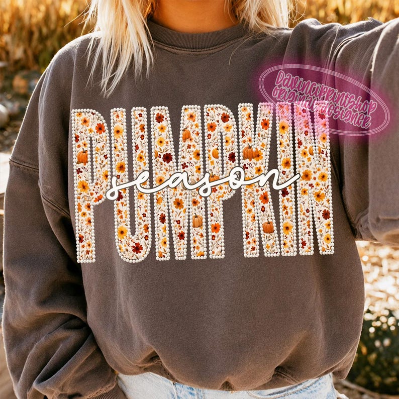 May include: A dark gray crewneck sweatshirt featuring the word PUMPKIN in large block letters filled with a floral and pumpkin pattern, overlaid with the word season in white cursive script. The block letters are outlined with white dots.