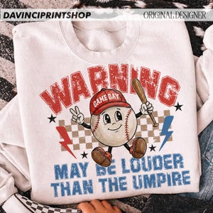 K&ouml;nnte beinhalten: Wei&szlig;es Sweatshirt mit Baseball-Grafik. Das Design zeigt einen Baseball-Charakter mit roter Kappe und Schl&auml;ger, mit dem Text "WARNING GAME DAY MAY BE LOUDER THAN THE UMPIRE". Enth&auml;lt Blitze und Sterne.