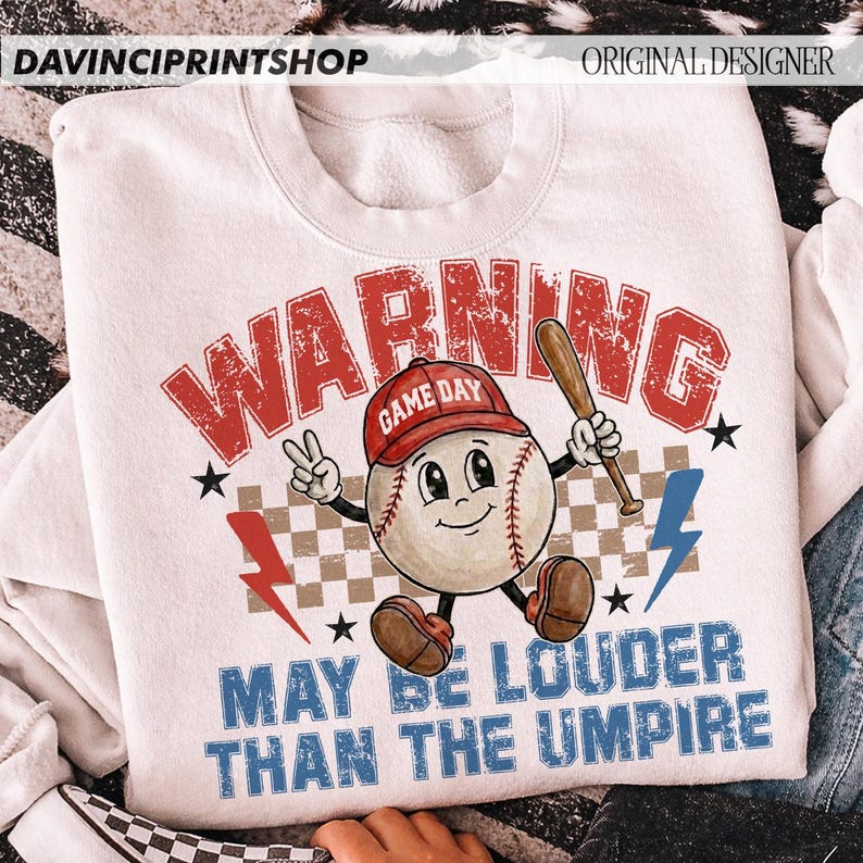 K&ouml;nnte beinhalten: Wei&szlig;es Sweatshirt mit Baseball-Grafik. Das Design zeigt einen Baseball-Charakter mit roter Kappe und Schl&auml;ger, mit dem Text "WARNING GAME DAY MAY BE LOUDER THAN THE UMPIRE". Enth&auml;lt Blitze und Sterne.