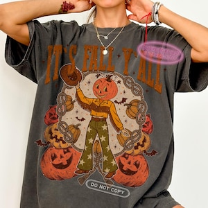 May include: Dark gray t-shirt with the text "IT'S FALL Y'ALL" in brown. The graphic features a pumpkin-headed figure in a cowboy hat and fringed shirt, surrounded by pumpkins and bats. The shirt also has the text "DO NOT COPY".