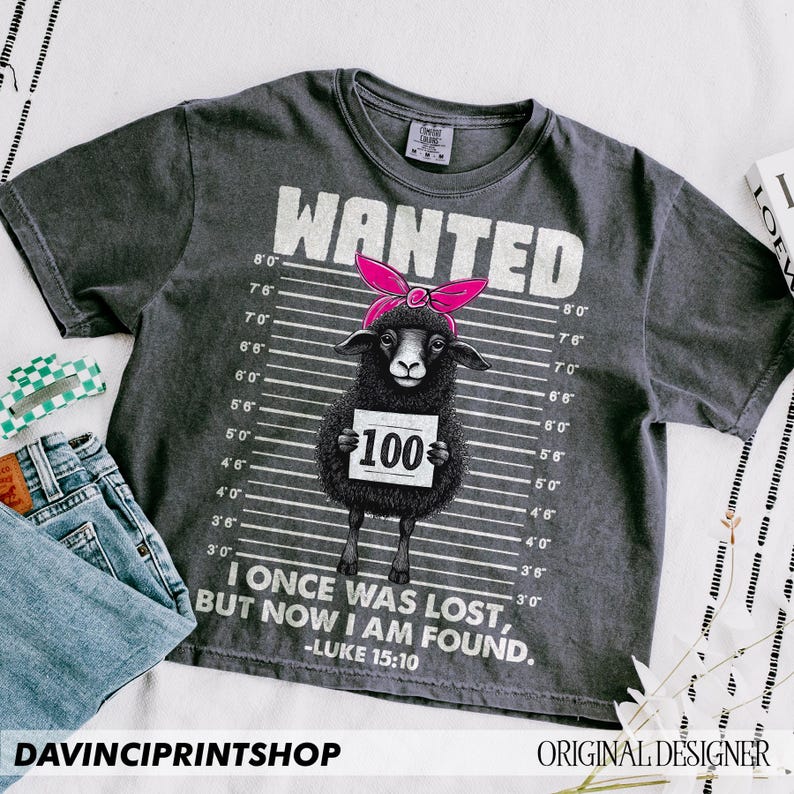 Puede incluir: Una camiseta gris oscuro con una oveja negra que lleva un pa&ntilde;uelo rosa y un cartel que dice "WANTED 100". La camiseta tiene un fondo blanco con l&iacute;neas negras y el texto "I once was lost, but now I am found. -Luke 15:10"