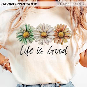 May include: A cream-coloured t-shirt displaying three daisy flowers in green, white, and orange, with yellow centres. The words "Life is Good" are written in black script below the flowers. The shirt has a crew neck.