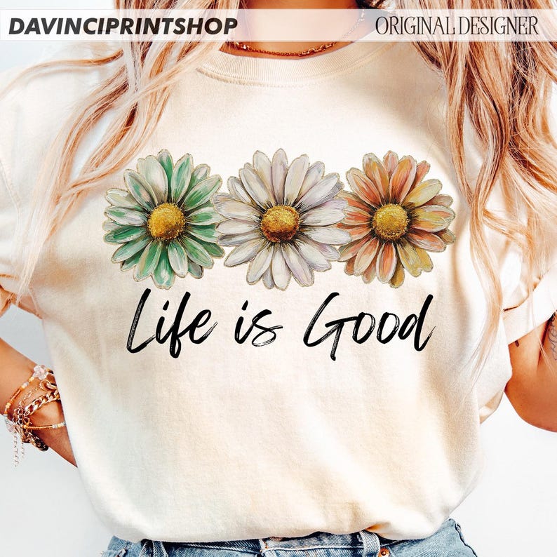 May include: A cream-coloured t-shirt displaying three daisy flowers in green, white, and orange, with yellow centres. The words "Life is Good" are written in black script below the flowers. The shirt has a crew neck.