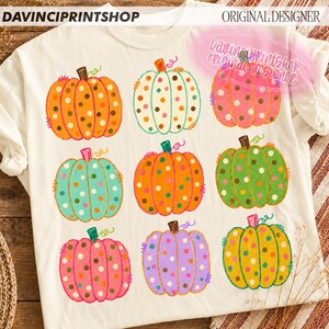 Puede incluir: Una camiseta de color crema con un estampado gr&aacute;fico de nueve calabazas de varios colores, incluyendo naranja, blanco, rosa, verde azulado, verde y morado. Cada calabaza est&aacute; decorada con lunares coloridos y tiene un peque&ntilde;o tallo verde.