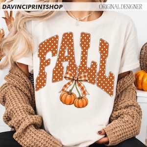 May include: A cream-colored short-sleeve t-shirt featuring the word FALL in large, orange, white-polka-dotted letters with a black outline. Below the text, two orange pumpkins are tied together with a brown and white plaid ribbon bow.