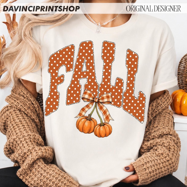 May include: A cream-colored short-sleeve t-shirt featuring the word FALL in large, orange, white-polka-dotted letters with a black outline. Below the text, two orange pumpkins are tied together with a brown and white plaid ribbon bow.