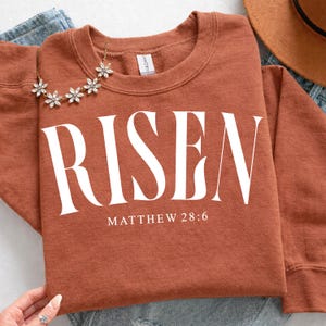 K&ouml;nnte beinhalten: Rostfarbenes Sweatshirt mit dem Wort "RISEN" in gro&szlig;en wei&szlig;en Buchstaben. Darunter steht der Text "MATTHEW 28:6". Eine goldfarbene Blumenkette liegt auf dem Sweatshirt.