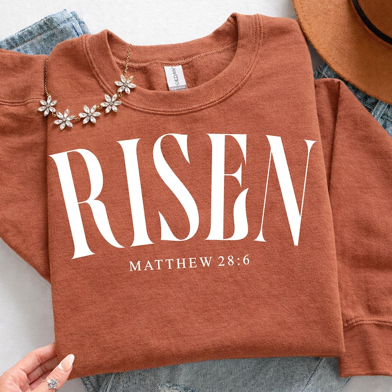 K&ouml;nnte beinhalten: Rostfarbenes Sweatshirt mit dem Wort "RISEN" in gro&szlig;en wei&szlig;en Buchstaben. Darunter steht der Text "MATTHEW 28:6". Eine goldfarbene Blumenkette liegt auf dem Sweatshirt.