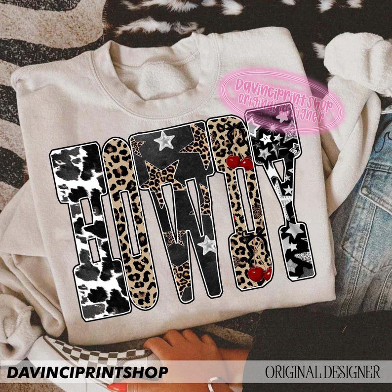 May include: Beige sweatshirt with the word "HOWDY" in a bold, decorative font. The letters feature cow print, leopard print, stars, and cherries. The text "DAVINCIPRINTSHOP ORIGINAL DESIGNER" is also visible.