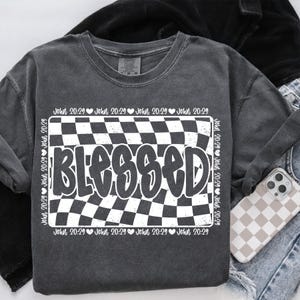 Op de afbeelding: Een donkergrijs T-shirt met een wit geruit patroon en het woord "BLESSED" in een vet, wit lettertype. Het geruite patroon wordt omringd door een rand met de tekst "John 20:29" die meerdere keren wordt herhaald.