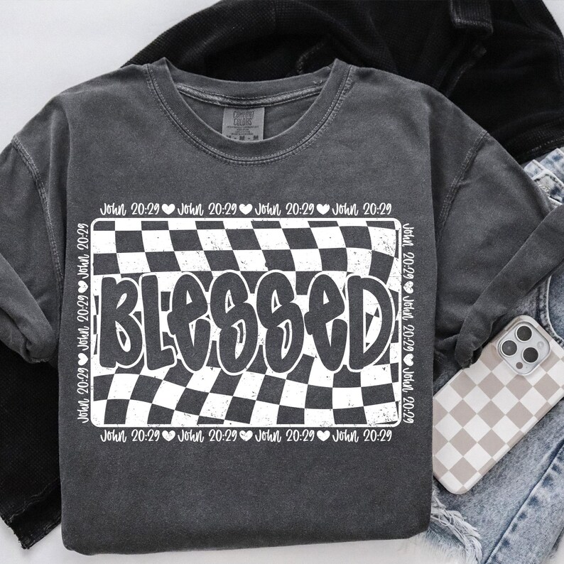 Op de afbeelding: Een donkergrijs T-shirt met een wit geruit patroon en het woord "BLESSED" in een vet, wit lettertype. Het geruite patroon wordt omringd door een rand met de tekst "John 20:29" die meerdere keren wordt herhaald.