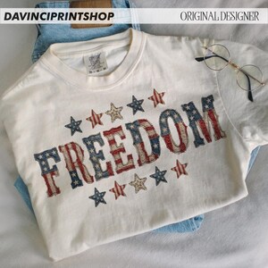 Pode incluir: Camiseta creme com a palavra "FREEDOM" em um design patri&oacute;tico desgastado. As letras s&atilde;o preenchidas com o padr&atilde;o da bandeira americana, cercadas por estrelas vermelhas, brancas e azuis. A camiseta est&aacute; dobrada sobre jeans azuis.