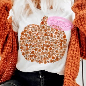 May include: White t-shirt featuring a pumpkin design made of orange and peach-colored flowers. The shirt has the text "Davinci print shop original designer" in a pink oval. The shirt is paired with a rust-colored sweater.