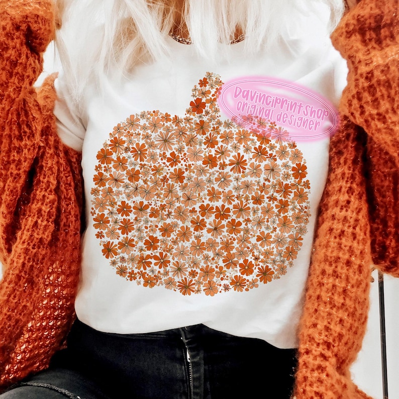 May include: White t-shirt featuring a pumpkin design made of orange and peach-colored flowers. The shirt has the text "Davinci print shop original designer" in a pink oval. The shirt is paired with a rust-colored sweater.