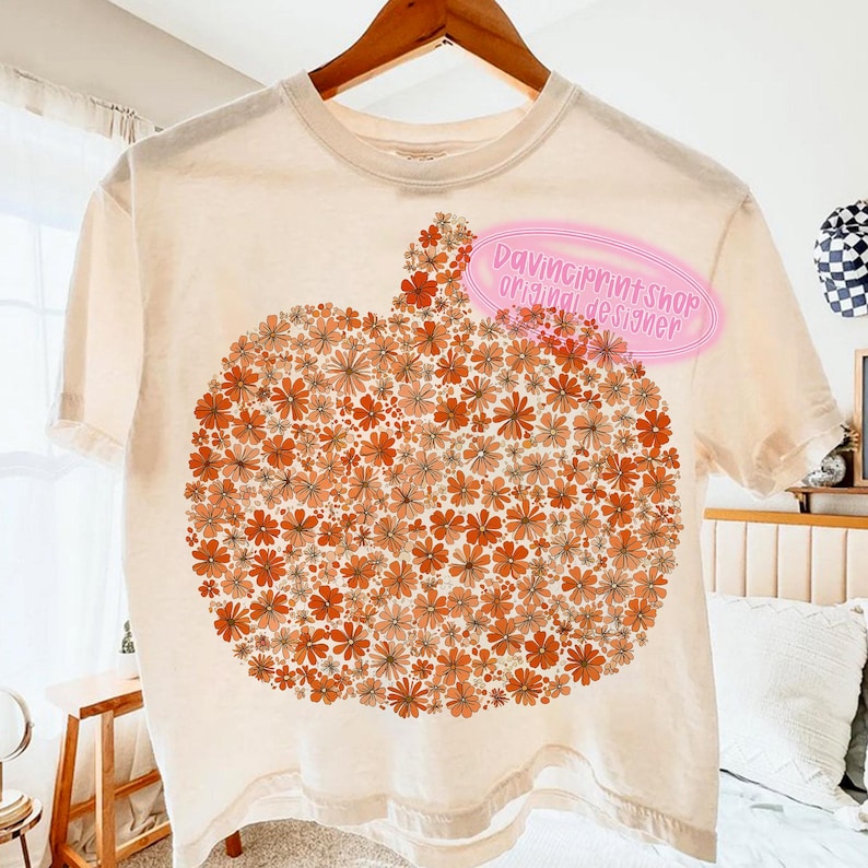 May include: Cream-colored t-shirt featuring a pumpkin design made of orange and peach-colored flowers. The shirt has short sleeves and a crew neck. The text "DavinciPrintShop original designer" is on the shirt.