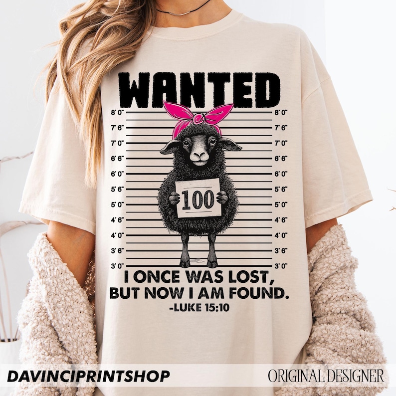 Puede incluir: Una camiseta beige con una ilustraci&oacute;n de una oveja negra que lleva una diadema rosa y sostiene un cartel que dice "100". La oveja est&aacute; en una alineaci&oacute;n policial con marcas de altura. El texto "WANTED" est&aacute; en la parte superior de la camiseta y el texto "I ONCE WAS LOST, BUT NOW I AM FOUND. -LUKE 15:10" est&aacute; en la parte inferior.