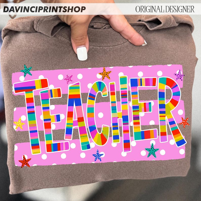 May include: A brown t-shirt featuring a rectangular pink graphic with white polka dots and the word TEACHER printed in large, multi-colored striped letters. The design is accented with small, colorful stars scattered across the pink background.