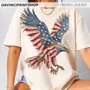 Pu&ograve; includere: T-shirt color crema con un design dettagliato di un'aquila. Le ali dell'aquila sono realizzate con toppe di denim blu con stelle bianche e strisce rosse e bianche, che ricordano la bandiera americana. Il testo "DAVINCIPRINTSHOP" e "ORIGINAL DESIGNER" sono visibili.