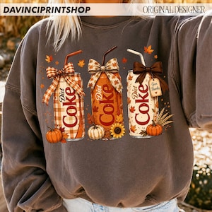 May include: A charcoal gray crewneck sweatshirt featuring a graphic of three Diet Coke cans decorated for autumn. The cans are adorned with bows, straws, pumpkins, sunflowers, and fall leaves. One can has a tag that reads Give Thanks.