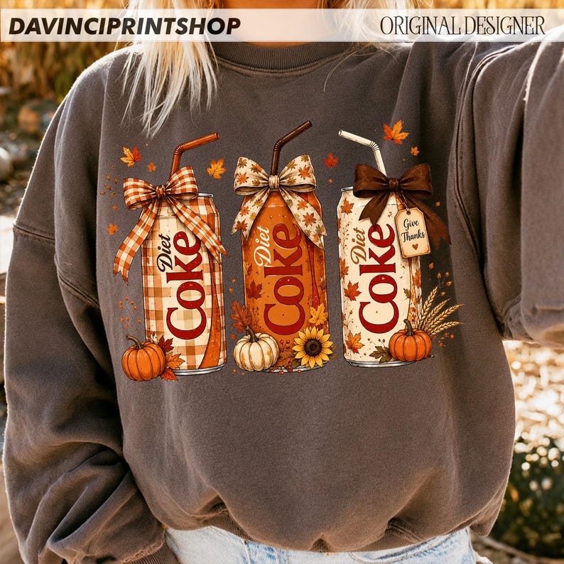 May include: A charcoal gray crewneck sweatshirt featuring a graphic of three Diet Coke cans decorated for autumn. The cans are adorned with bows, straws, pumpkins, sunflowers, and fall leaves. One can has a tag that reads Give Thanks.