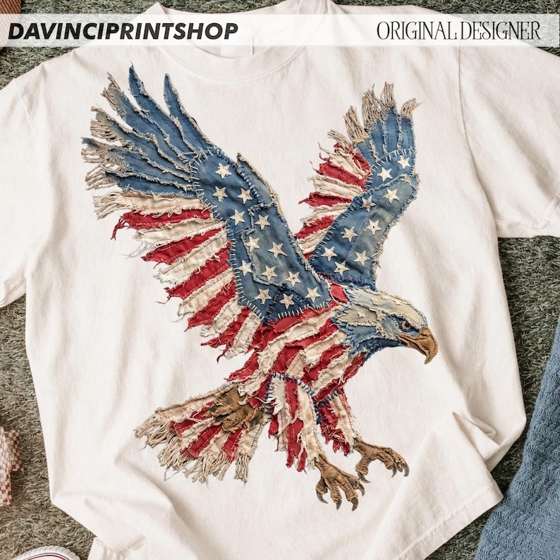 Pu&ograve; includere: T-shirt bianca con un design patriottico di un'aquila calva americana. L'aquila &egrave; composta da toppe rosse, bianche e blu, che ricordano la bandiera americana. I testi "DAVINCIPRINTSHOP" e "ORIGINAL DESIGNER" sono visibili.