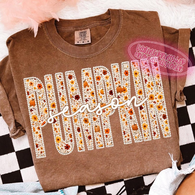 May include: A brown short-sleeve t-shirt featuring the word PUMPKIN in large block letters filled with a floral and pumpkin pattern, outlined in white dots. The word season is written in white cursive script across the center of the design.