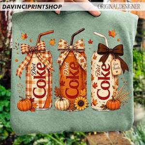 May include: A sage green sweatshirt featuring a graphic of three Diet Coke cans decorated with autumn themes. Each can has a straw, a bow, and fall elements like pumpkins, sunflowers, and leaves. One can has a tag that reads Give Thanks.