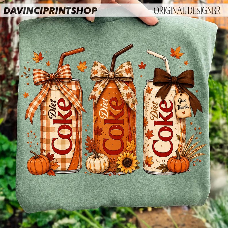 May include: A sage green sweatshirt featuring a graphic of three Diet Coke cans decorated with autumn themes. Each can has a straw, a bow, and fall elements like pumpkins, sunflowers, and leaves. One can has a tag that reads Give Thanks.