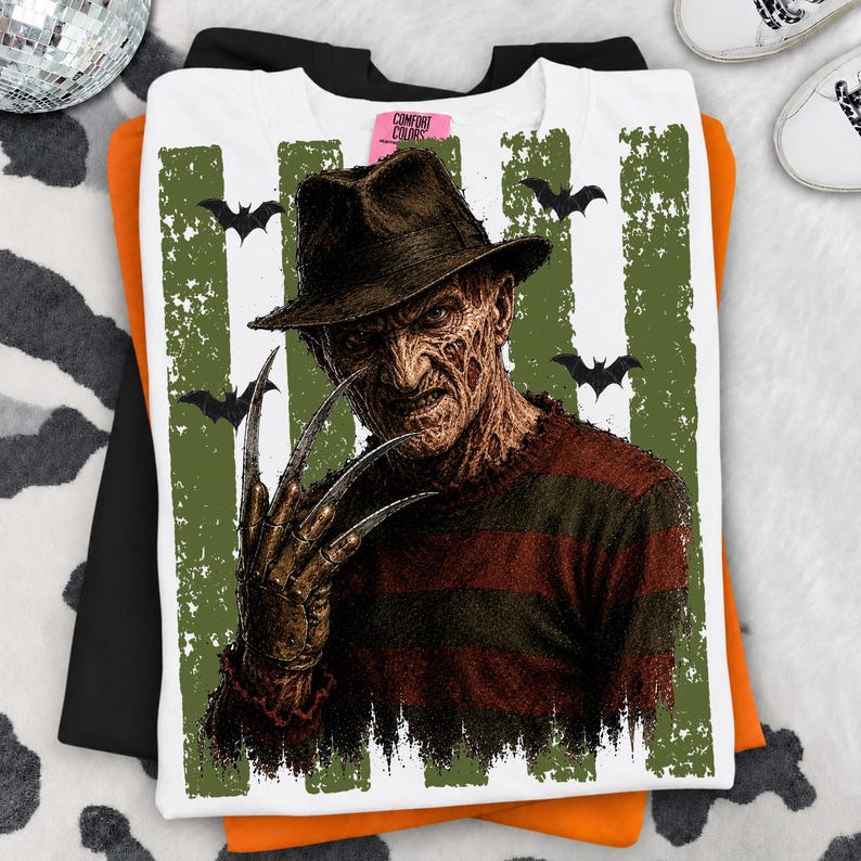 May include: A white t-shirt featuring a graphic print of a character wearing a brown fedora and a red and dark brown striped sweater. The character has a scarred face and a metal clawed glove. Green vertical distressed stripes and bats are in the background.