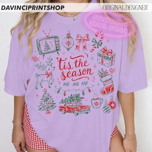 May include: A light purple t-shirt featuring a festive red and green graphic print with the text 'tis the season' and 'HO HO HO'. The design includes a vintage television, a reindeer, a Christmas tree, a car, a gift, and a stocking.