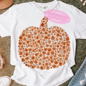 May include: White t-shirt featuring a pumpkin design made of orange and peach-colored flowers. The shirt has a crew neck and short sleeves. A pink tag reads "DaVinciPrintShop original designer."