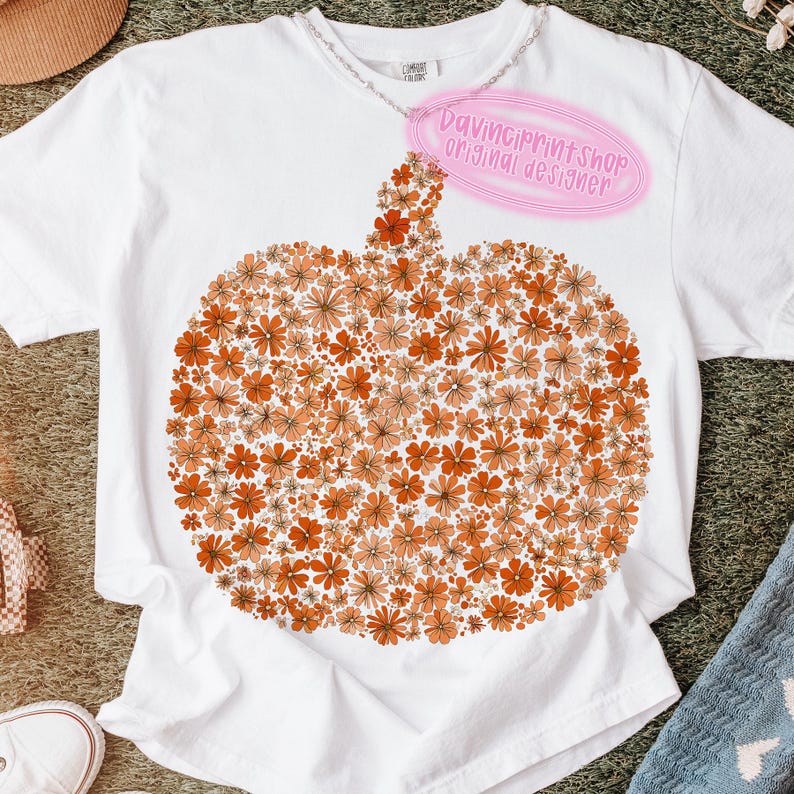 May include: White t-shirt featuring a pumpkin design made of orange and peach-colored flowers. The shirt has a crew neck and short sleeves. A pink tag reads "DaVinciPrintShop original designer."
