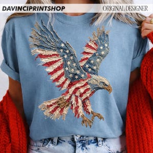 Pu&ograve; includere: Una maglietta azzurra con un design di aquila della bandiera americana invecchiata. Le ali dell'aquila sono spiegate, mostrando le stelle e le strisce in uno stile vintage e strutturato. La maglietta ha le maniche arrotolate e il testo "DAVINCIPRINTSHOP" e "ORIGINAL DESIGNER".