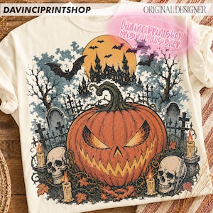 May include: An off-white t-shirt featuring a graphic print of a large orange jack-o'-lantern, two human skulls, and lit candles in a graveyard setting. The background shows a dark castle, a full moon, bats, and bare trees under a cloudy sky.