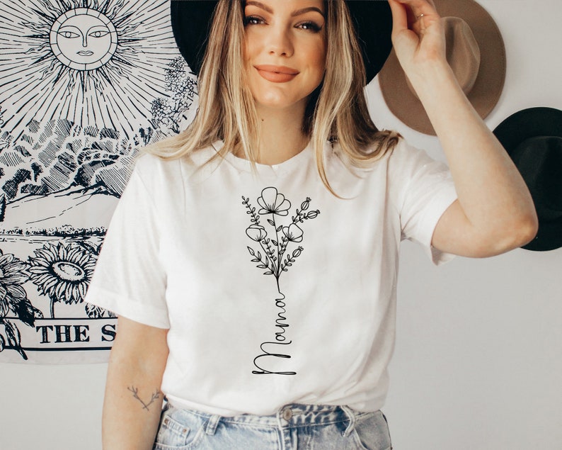 Pode incluir: Camiseta branca com um desenho de flores em linha preta e a palavra "Mama" escrita verticalmente.
