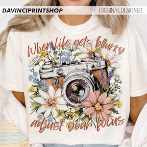 May include: Cream-colored t-shirt featuring a vintage camera design surrounded by colorful flowers. The text "When life gets blurry, adjust your focus" is written in a script font. The shirt has the text "DAVINCIPRINTSHOP" and "ORIGINAL DESIGNER" at the top.