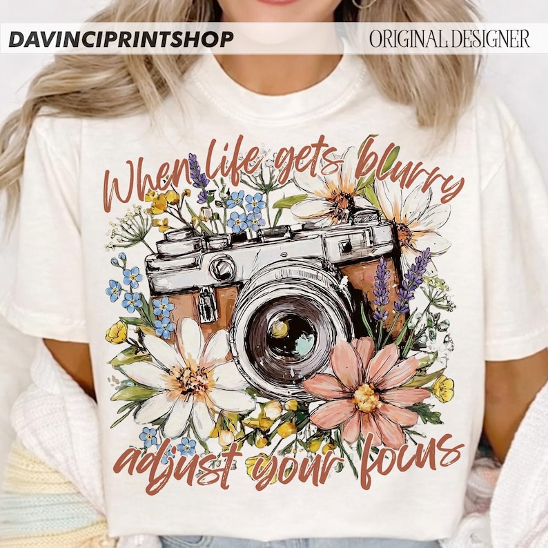 May include: Cream-colored t-shirt featuring a vintage camera design surrounded by colorful flowers. The text "When life gets blurry, adjust your focus" is written in a script font. The shirt has the text "DAVINCIPRINTSHOP" and "ORIGINAL DESIGNER" at the top.