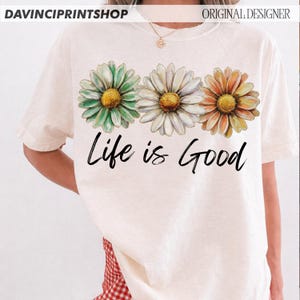 May include: Cream-coloured t-shirt featuring three daisy flowers in green, white, and orange with yellow centres. The phrase "Life is Good" is written in black script below the flowers. The shirt has short sleeves.