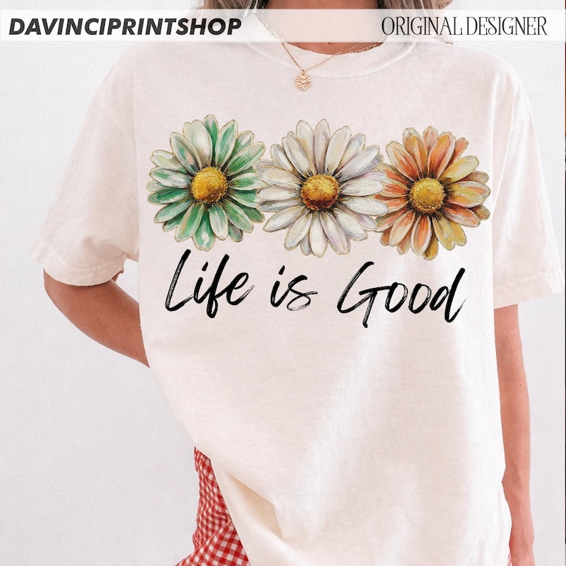 May include: Cream-coloured t-shirt featuring three daisy flowers in green, white, and orange with yellow centres. The phrase "Life is Good" is written in black script below the flowers. The shirt has short sleeves.