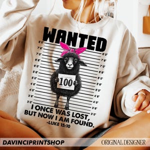 Puede incluir: Una sudadera blanca con un gr&aacute;fico en blanco y negro de una oveja negra con un lazo rosa. El gr&aacute;fico est&aacute; en estilo de cartel de b&uacute;squeda con una tabla de altura y el texto "WANTED" en la parte superior. La oveja sostiene un cartel que dice "100". El texto "I ONCE WAS LOST, BUT NOW I AM FOUND." est&aacute; debajo del gr&aacute;fico, con la referencia b&iacute;blica "LUKE 15:10" debajo.