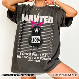 Puede incluir: Una camiseta gris con una oveja negra que lleva un pa&ntilde;uelo rosa y sostiene un cartel que dice "100 &euro;". La camiseta tiene un dise&ntilde;o de cartel de b&uacute;squeda con medidas de altura y el texto "WANTED" en la parte superior. El texto "Estaba perdido, pero ahora he sido encontrado." est&aacute; impreso debajo de la oveja, junto con la referencia b&iacute;blica "Lucas 15:10".