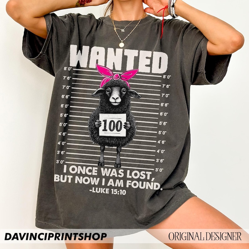 Puede incluir: Una camiseta gris con una oveja negra que lleva un pa&ntilde;uelo rosa y sostiene un cartel que dice "100 &euro;". La camiseta tiene un dise&ntilde;o de cartel de b&uacute;squeda con medidas de altura y el texto "WANTED" en la parte superior. El texto "Estaba perdido, pero ahora he sido encontrado." est&aacute; impreso debajo de la oveja, junto con la referencia b&iacute;blica "Lucas 15:10".